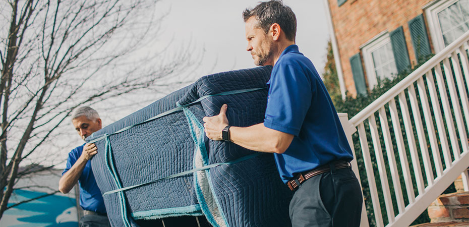 Two movers are carrying a padded piece of furniture out of a house.