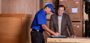 A man in a suit discusses with a mover in a warehouse with crates in the background.
