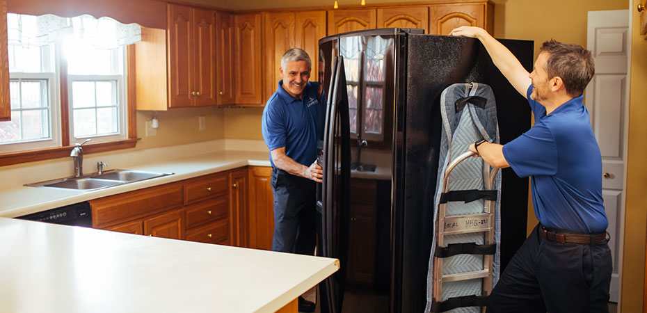 Two movers move a refrigerator out of a kitchen with a dolly.