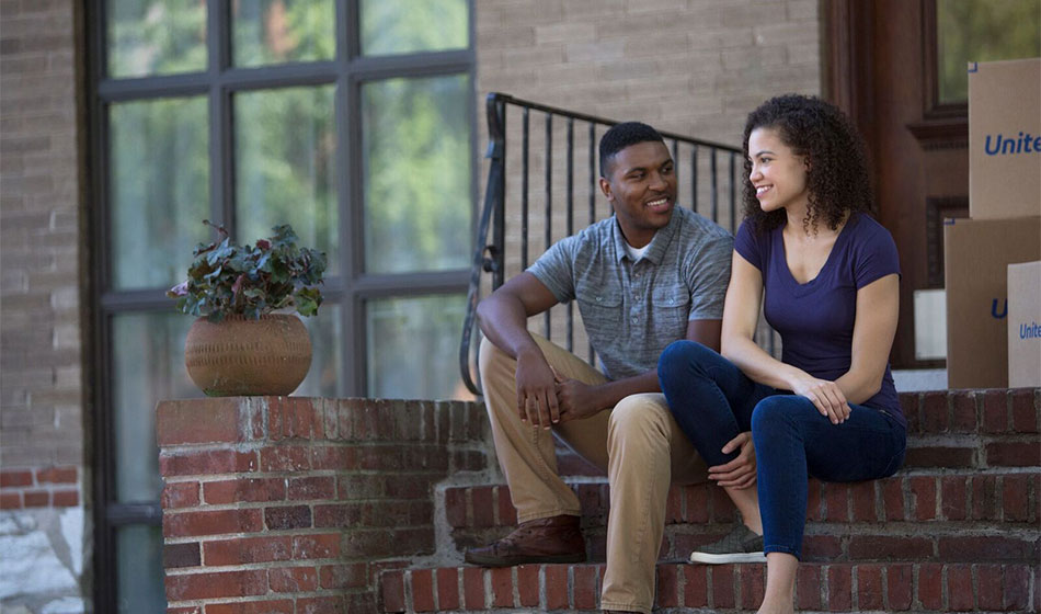 A couple sits on the front steps of their house with boxes behind them.