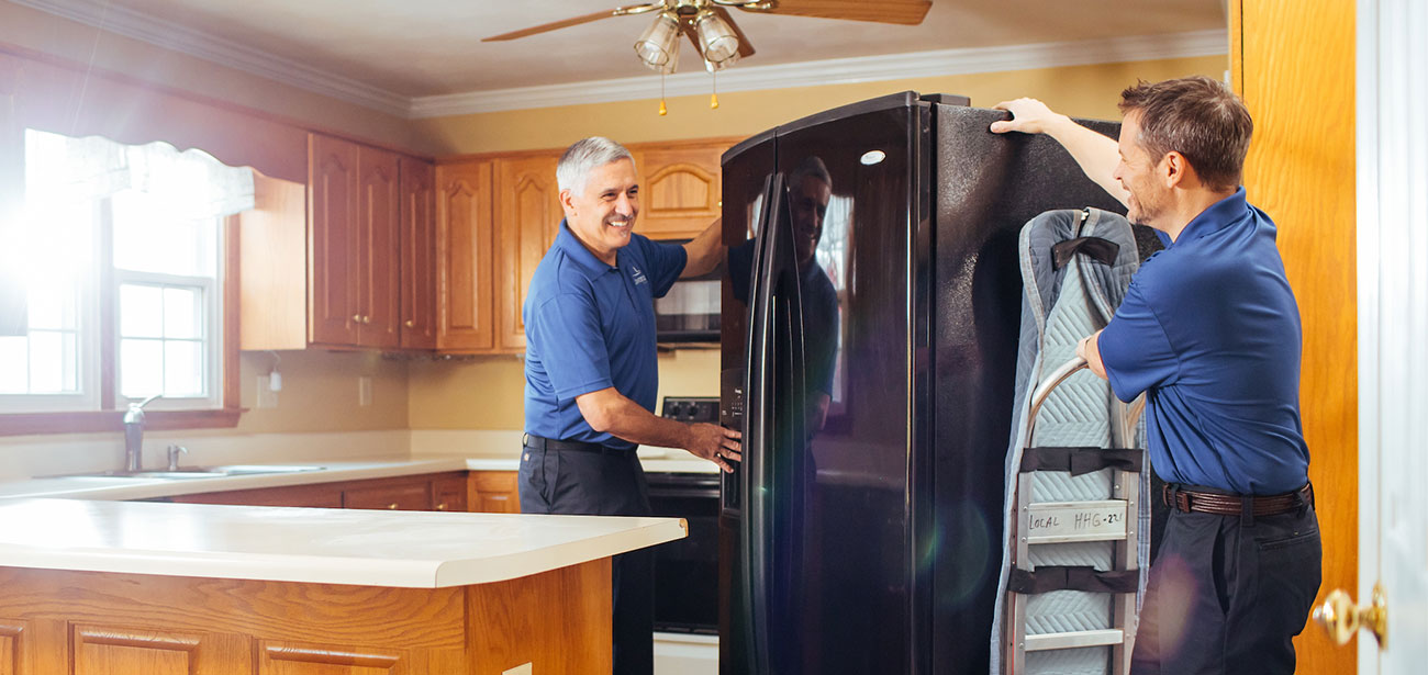 Two movers move a fridge out of a kitchen with a dolly.