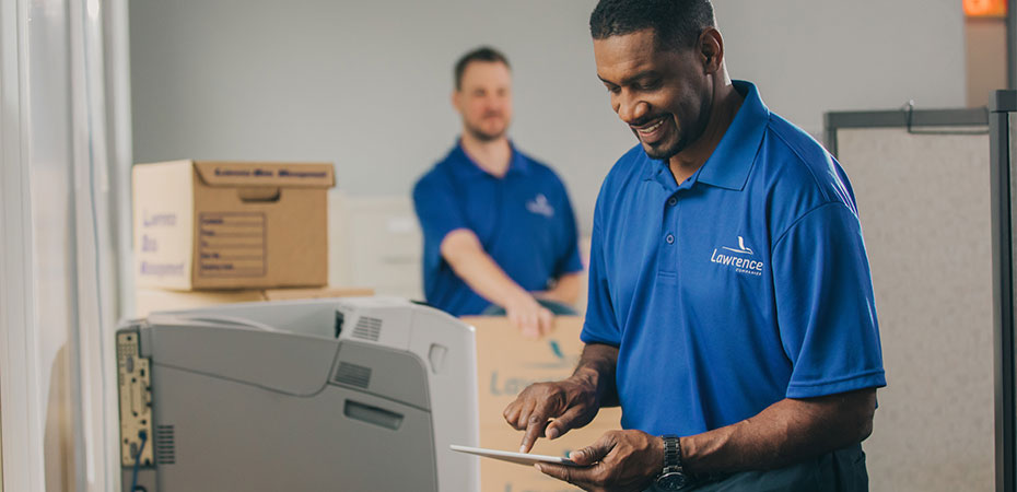 A mover holding a tablet with another mover unboxing in the background.