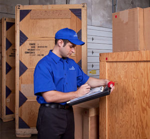 A commercial mover in a warehouse standing in front of wooden crate checking off a list.