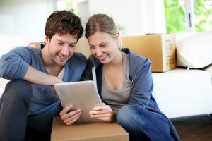 Woman and man sitting on floor looking a tablet with moving boxes in the background