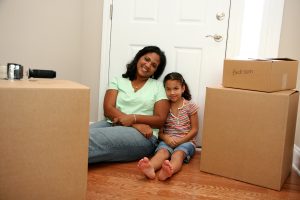 Woman and young girl sitting on the floor of bedroom leaning against the door with moving boxes staked around them.