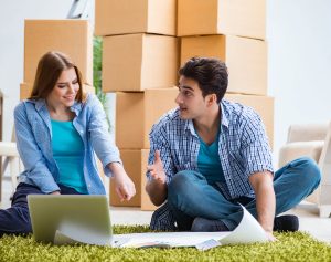 woman and man sitting on the ground with stacked moving boxes behind them and a laptop and papers in front of them as they plan a move.