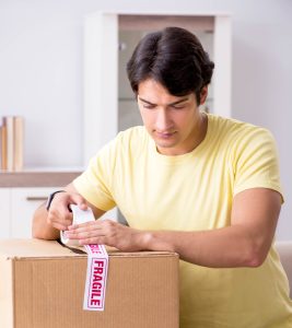 Man in yellow shirt taping closed a moving carton with tape that says fragile.