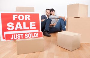 Man an woman sit on wooden house floor in the background with moving boxes and For Sale Just Sold sign in the foreground.