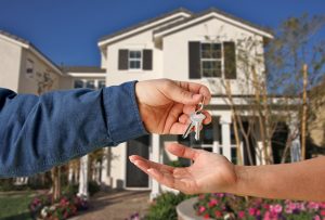 House keys being exchanged from one hand to another with a house in the background.