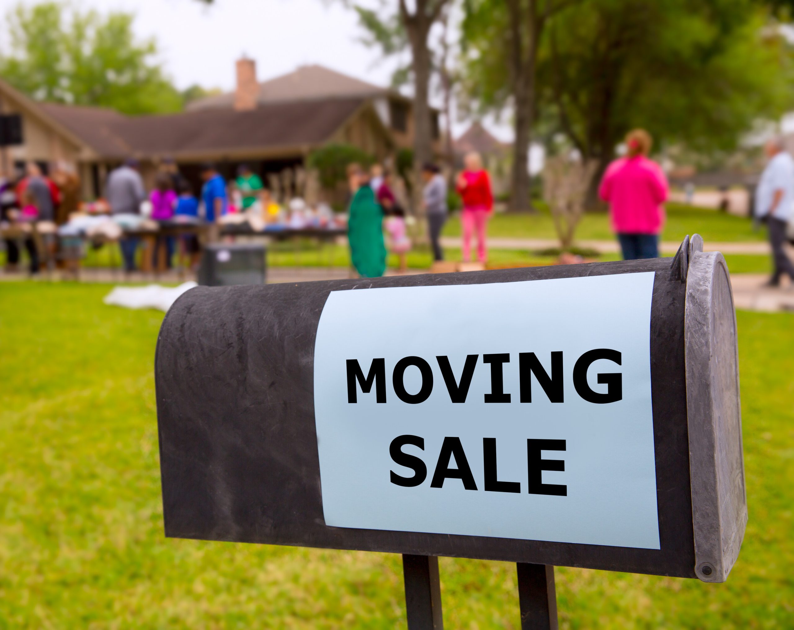 Moving sale sign posted on a mailbox with people in the background out of focus shopping at a moving sale.