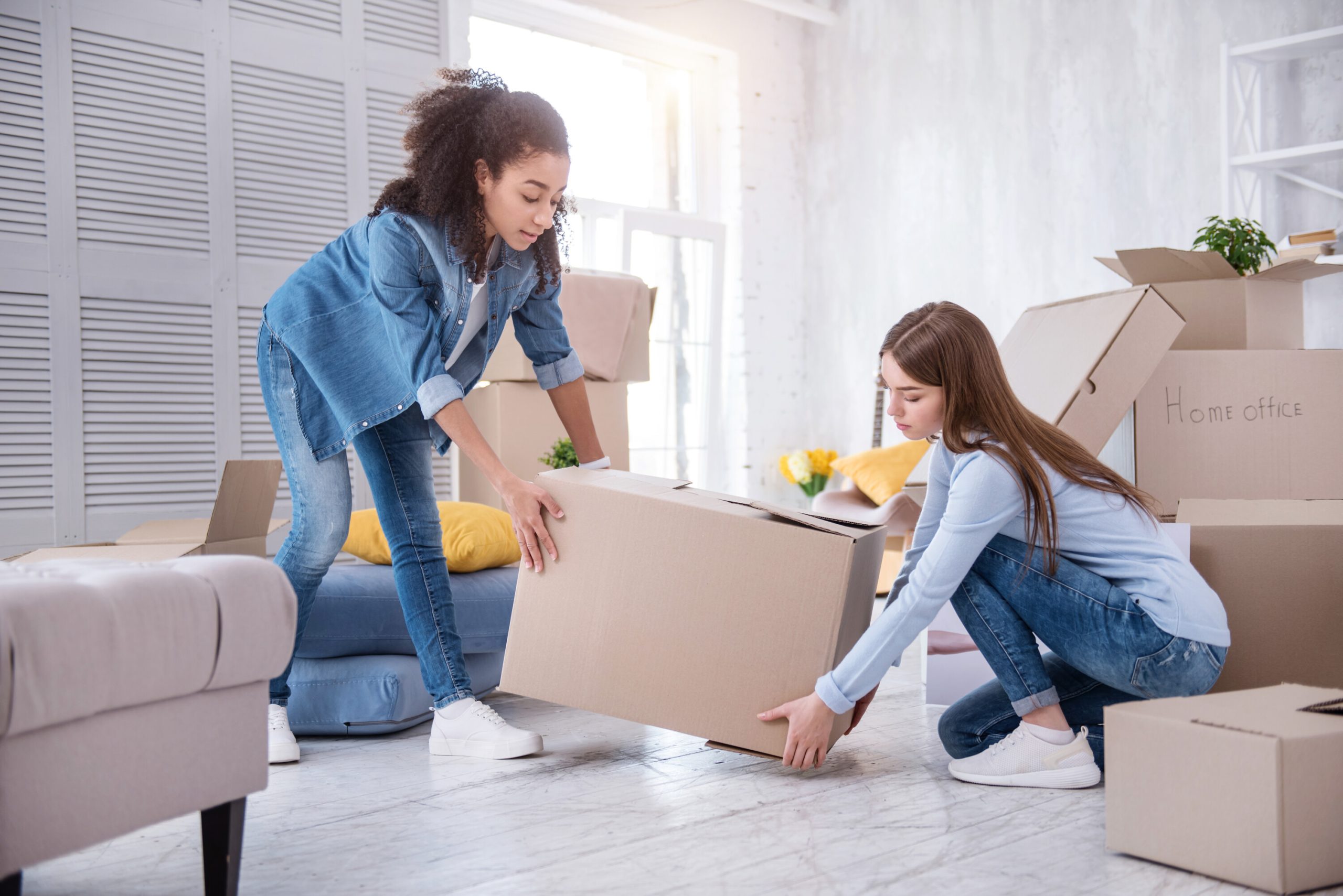 Two young women with long brown hair and dressed in jeans lift a moving box in a living room with numerous other moving boxes in the background.