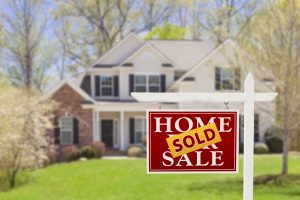 For sale sign with a sold sticker on in in a yard of a two story white and brick house.