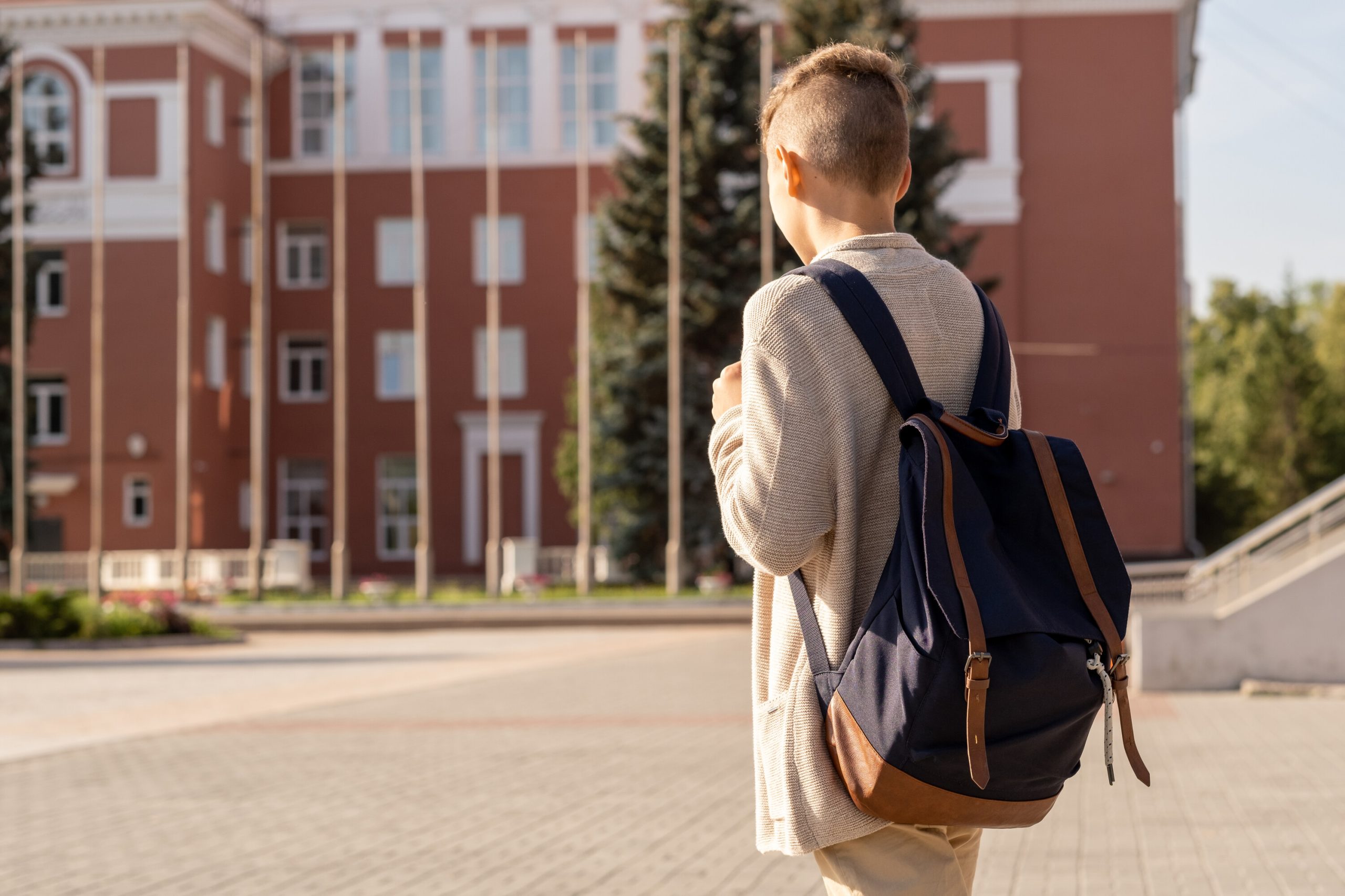 Youthful boy with a black backpack stands in front of a new school.
