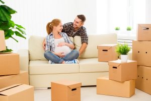 Pregnant woman sitting on couch talking to man behind couch while surrounded by moving boxes.