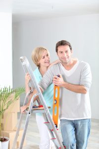 Man and woman near ladder holding a ruler with moving boxes in the background.