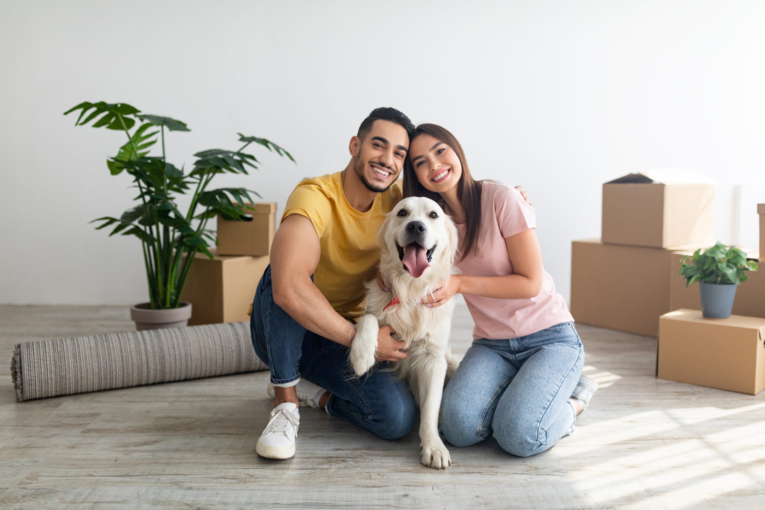 Cheery young couple with cute golden retriever dog sitting on floor of new home on relocation day. Happy millennial husband and wife with cute pet moving to new house