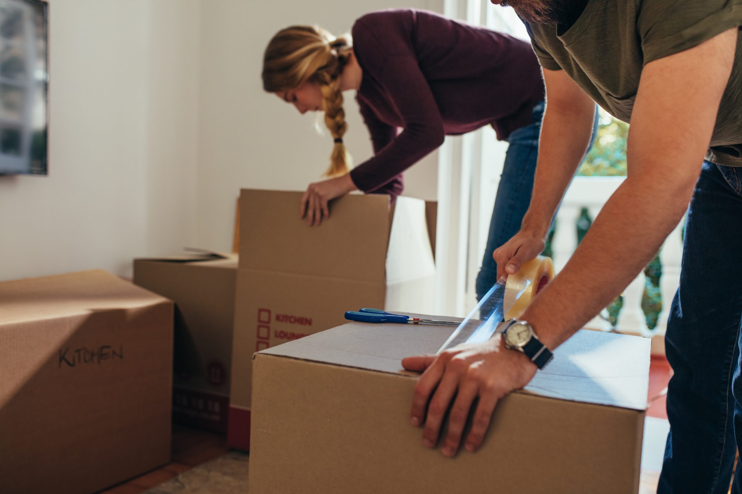 Close up of man sealing a packing box with adhesive tape while a woman placing articles in another box. Couple packing their items for moving into a new house.