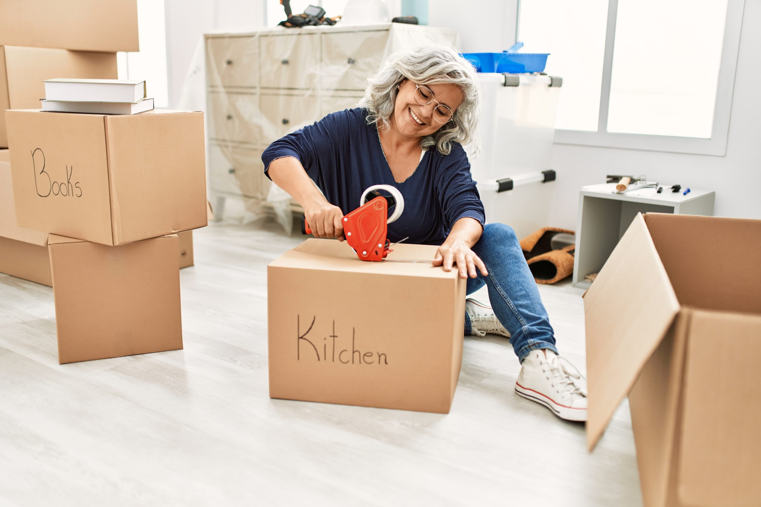 Middle age grey-haired woman smiling happy packing kitchen cardboard box at new home.