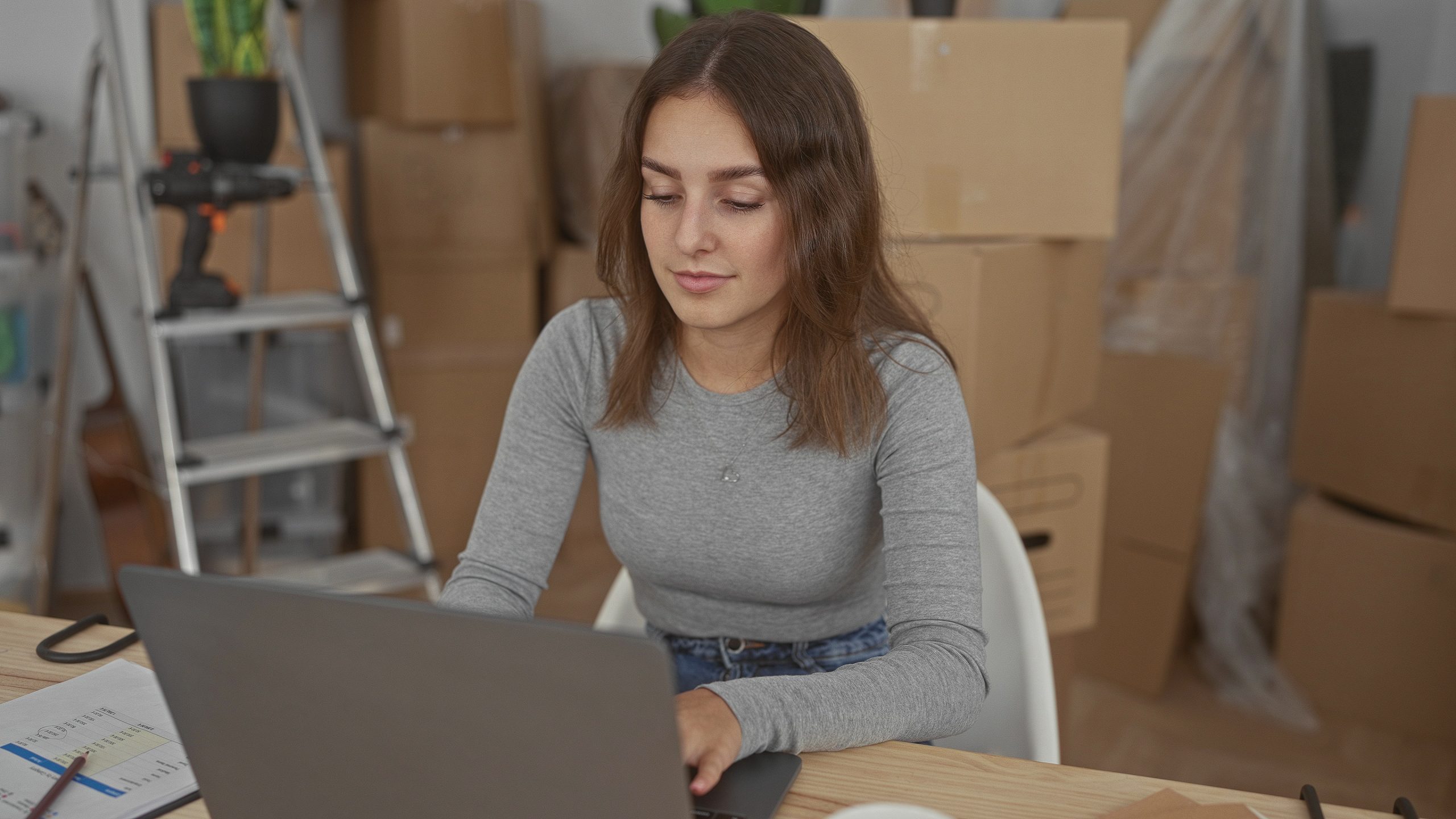 Woman typing on laptop changing address in building with stacked moving boxes and ladder