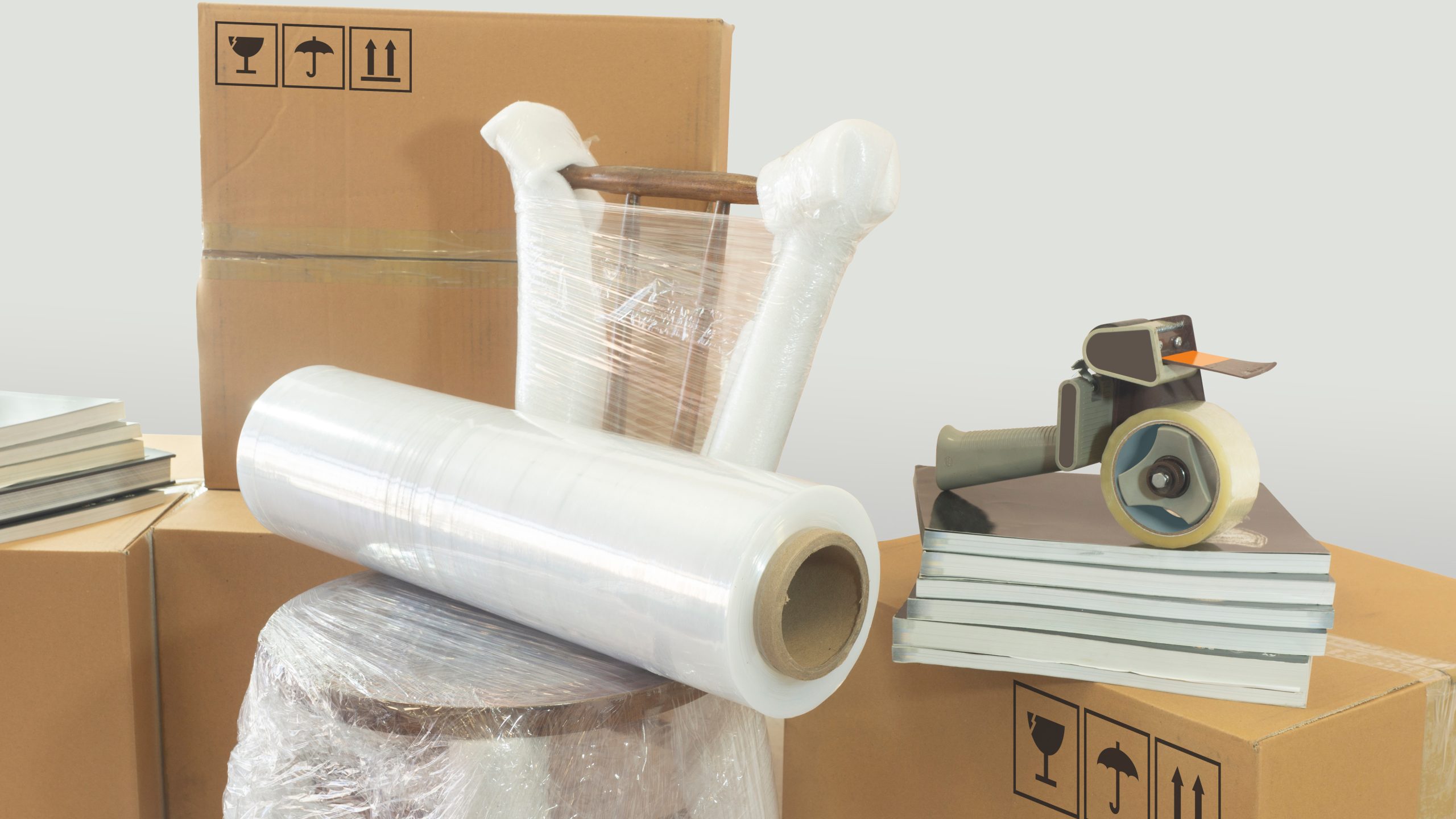 Moving scene with a roll of plastic to pack on a chair wrapped in plastic with closed cardboard boxes, books and packing tape on white background